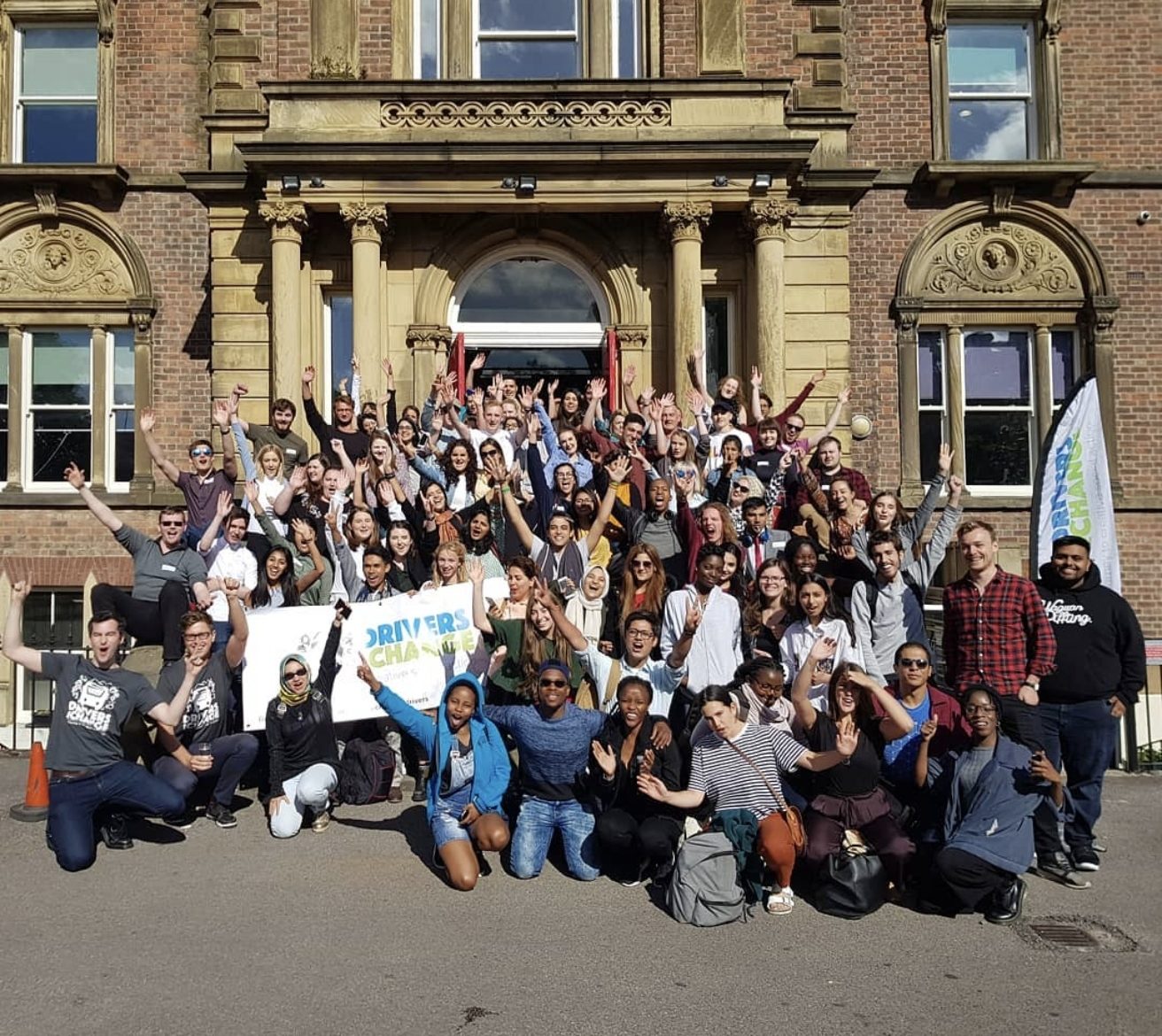 The Drivers for Change cohort gathered outside a building, arms raised, with the programme banner